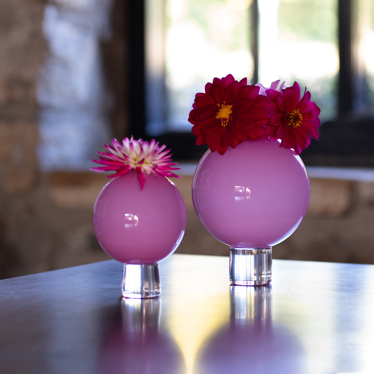 Two pink glass posey vases displayed on a reflective tabletop with dahlia flowers placed on top, against a softly blurred indoor background.