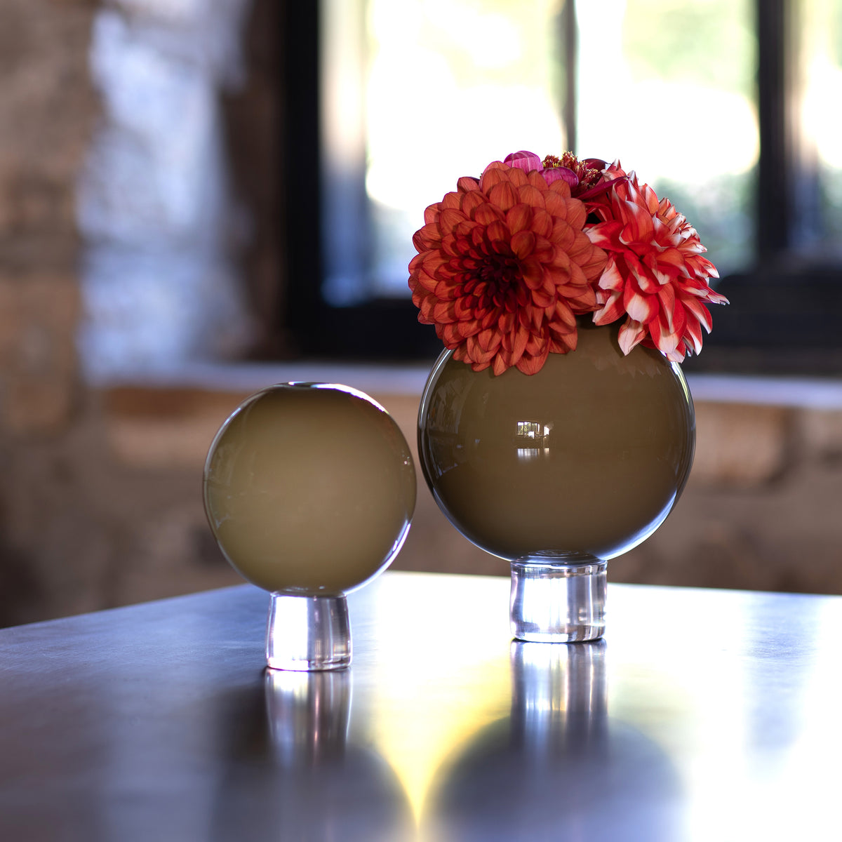 Two round glass hand blown vases, one small and one large, filled with orange and red dahlias