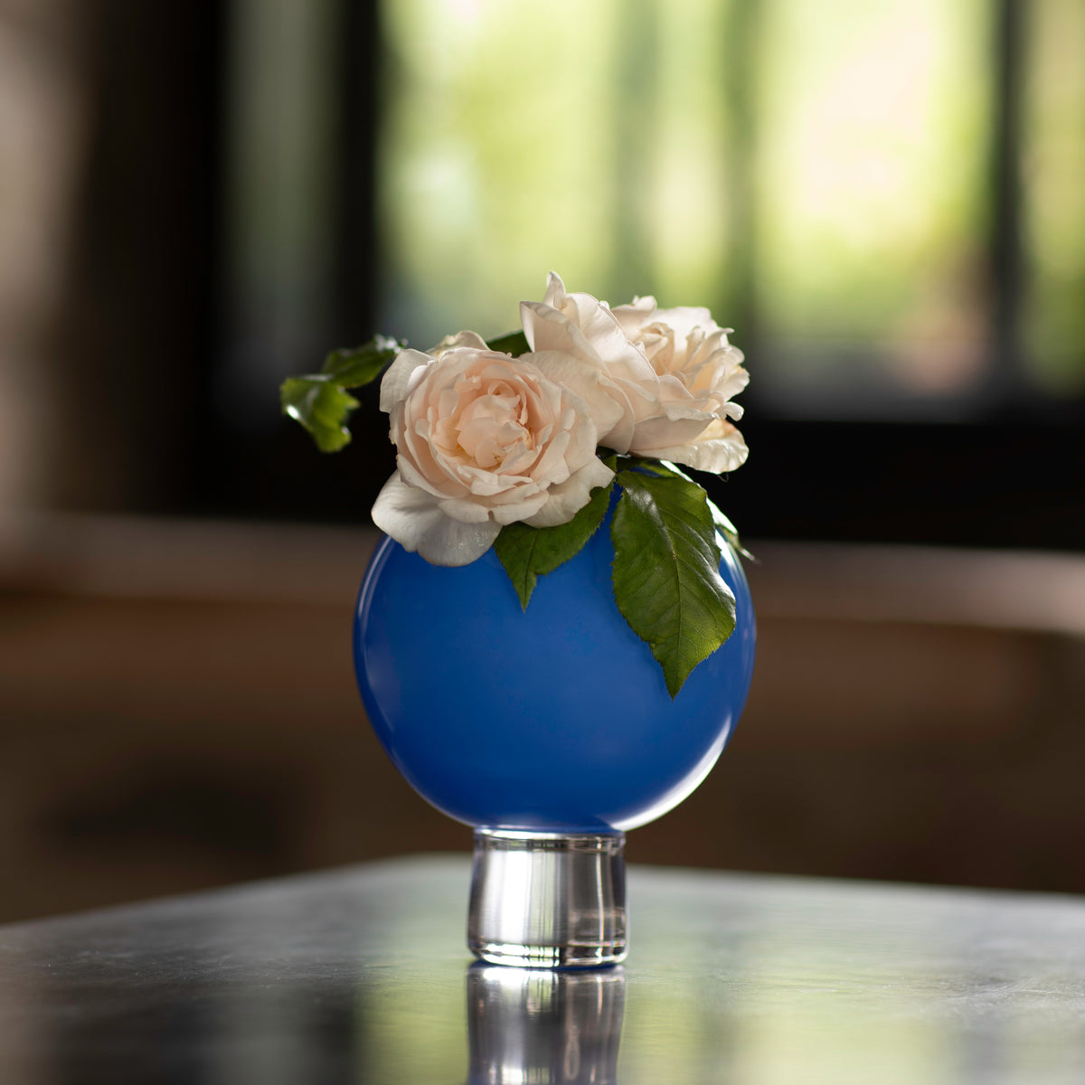 A small blue glass ball vase on a table, filled with water and containing a small posey of beige and pink flowers.