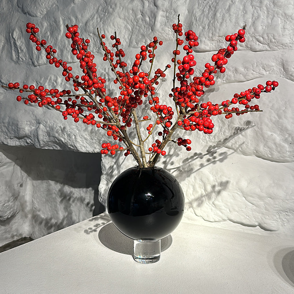 a round black glass vase on a table with a display of Christmas Ilex