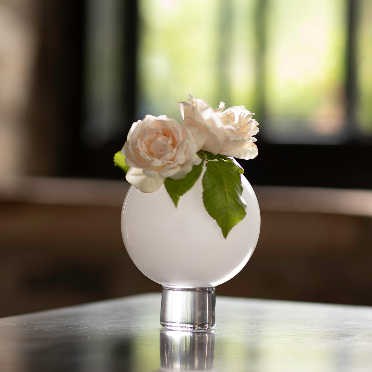 A small white ball vase with a single bloom of a rose with green leaves placed on a reflective surface.