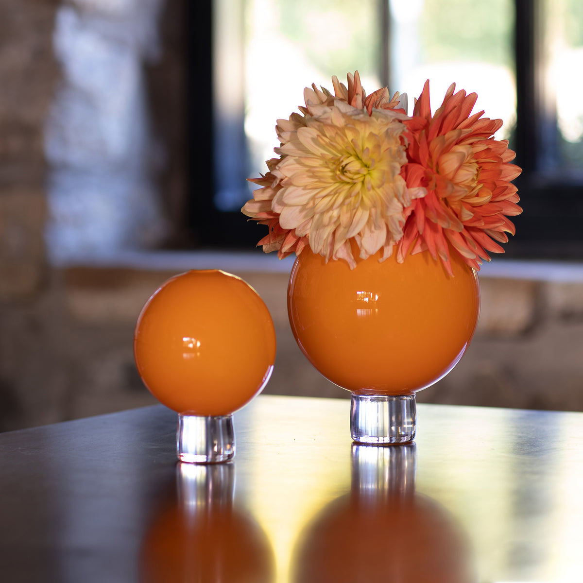 Two orange decorative round vases on clear glass bases, with the larger sphere holding a bouquet of orange and cream dahlias