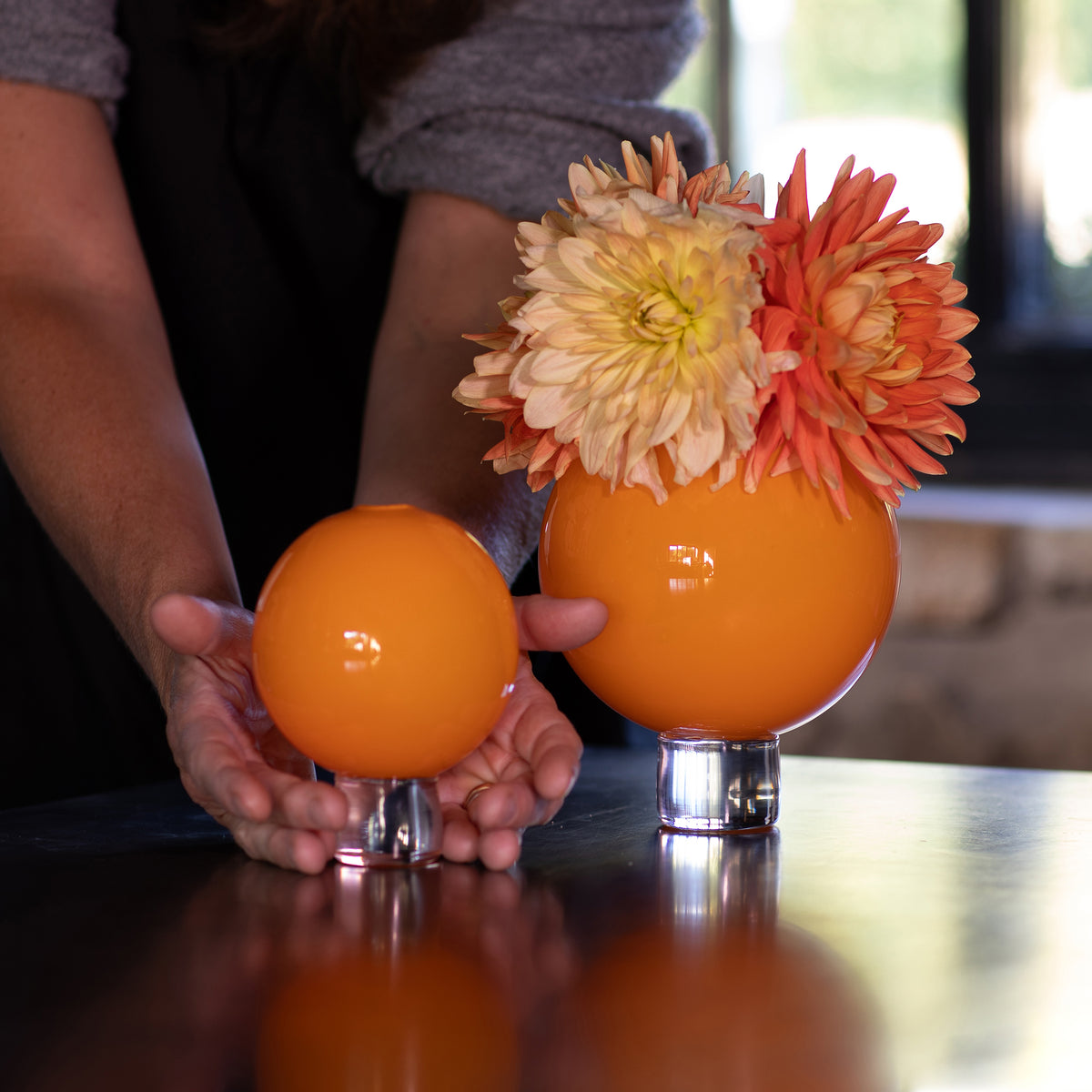 two hand blown Halloween orange round vases, with hands for scale and Dahlia flowers