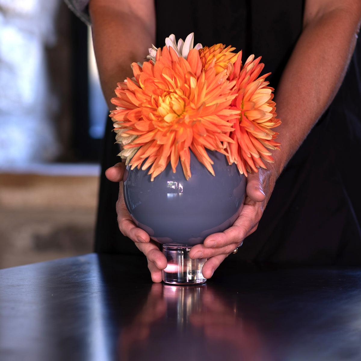 A person holds a round gray glass pedestal vase filled with large orange and yellow dahlia flowers on a dark reflective tabletop.