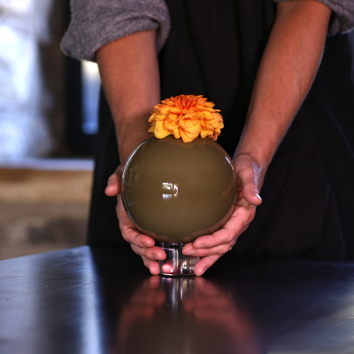 hands holing a large round, glossy olive-green glass vase on a small clear base, topped with an orange dahlia flower