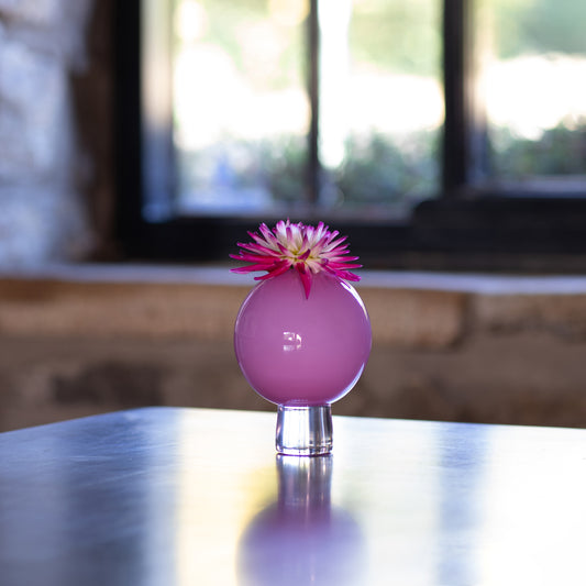 A pink glass orb vase with a clear glass pedestal base, holding a pink and purple dahlia flower, set on a reflective dark table with a blurred window background.