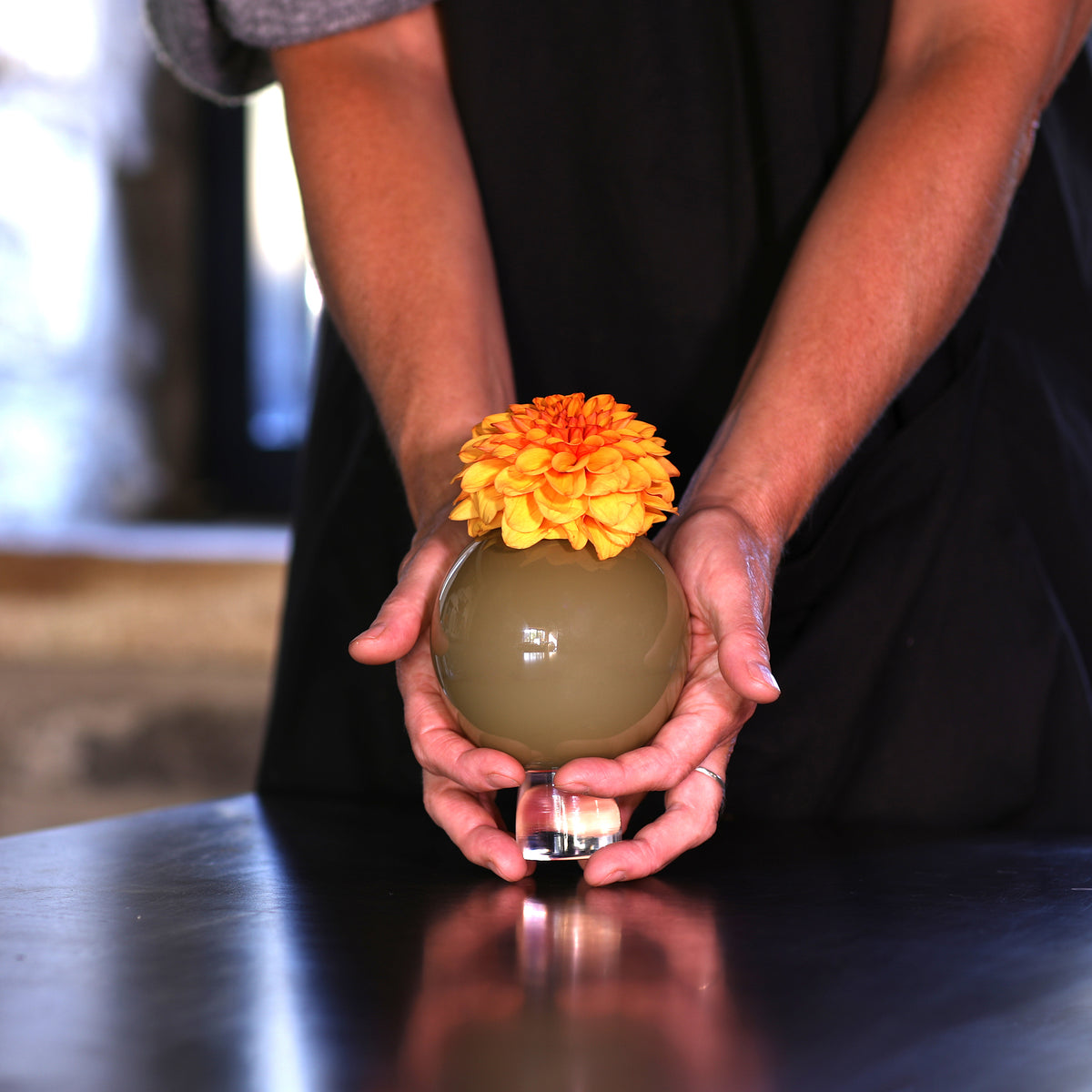 hands holding a small round glas  olive-green vase and topped with a bright orange dahlia, set on a dark tabletop.