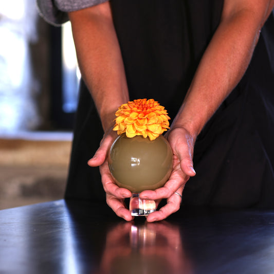 hands holding a small round glas  olive-green vase and topped with a bright orange dahlia, set on a dark tabletop.