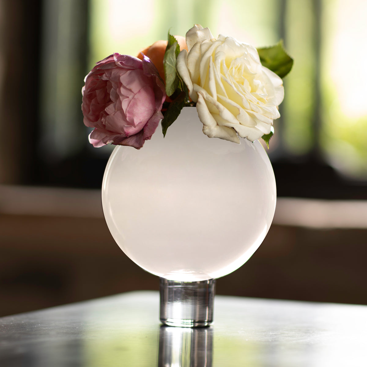 A white glass vase in the shape of a large ball, with a small hole on the top, partially filled with water and displaying two roses and a peony.